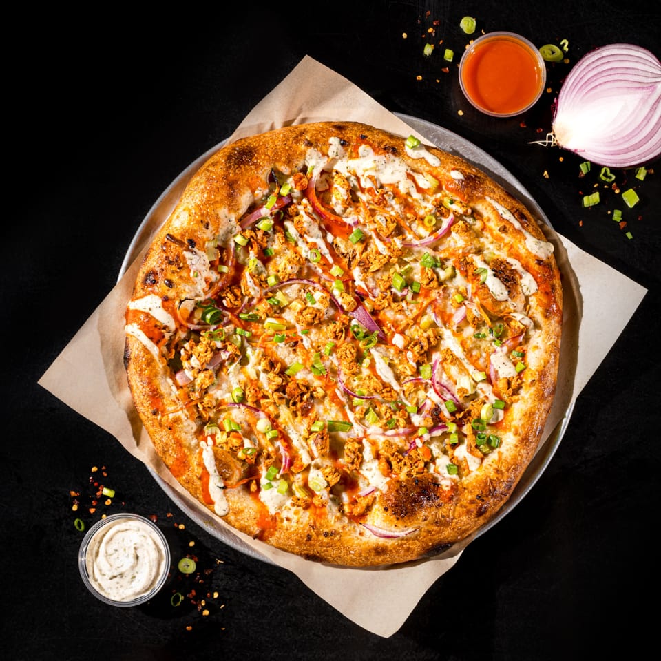 Vegan Buffalo Chicken pizza with red onions, green onions, and ranch drizzle on a dark surface.