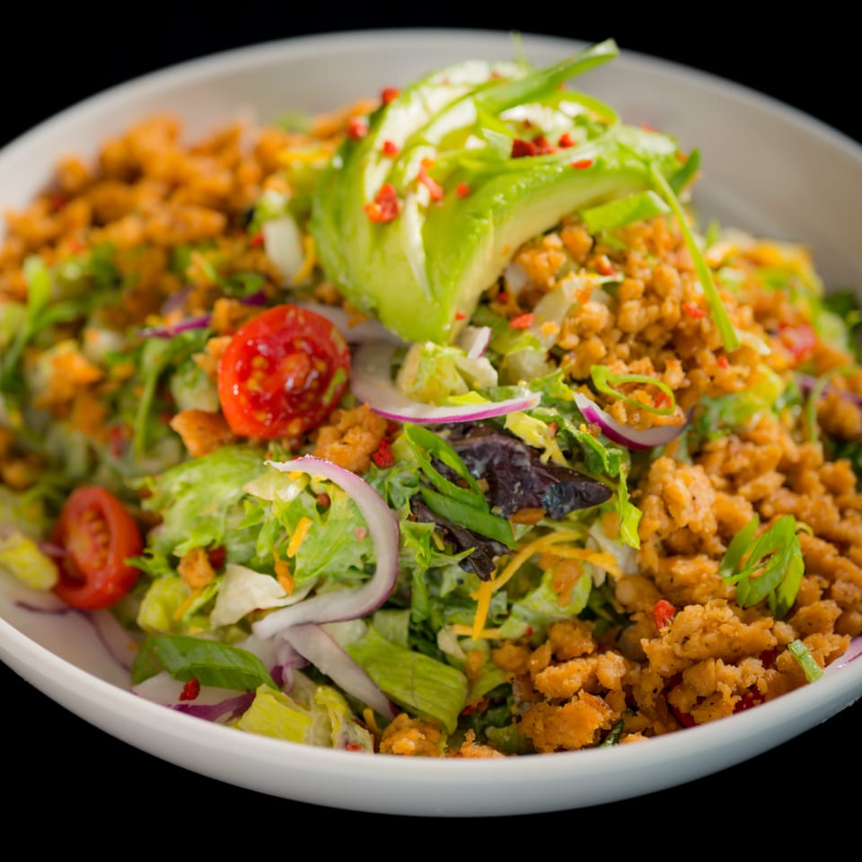 Vegan salad with lettuce, tomatoes, red onions, avocado, and plant-based crumbles in a white bowl.