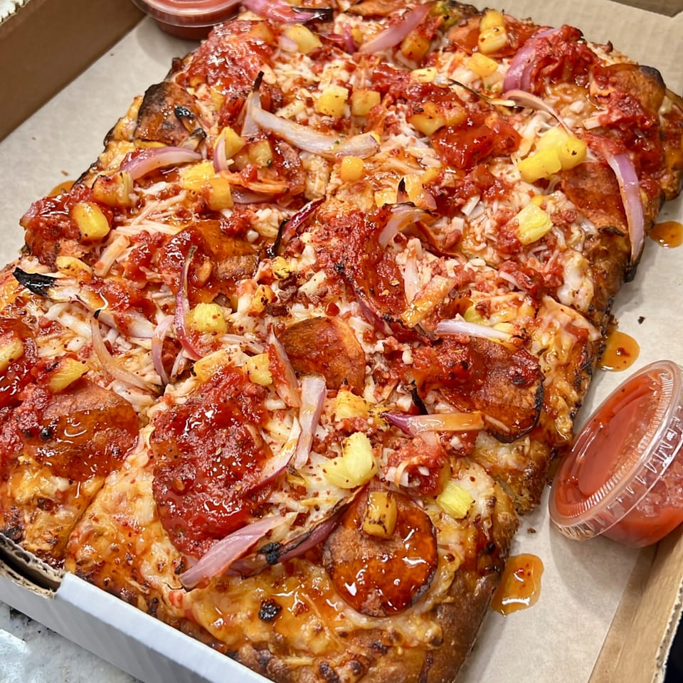 Vegan BBQ pizza with pineapple, red onion, and pepperoni in a pizza box with dipping sauce.