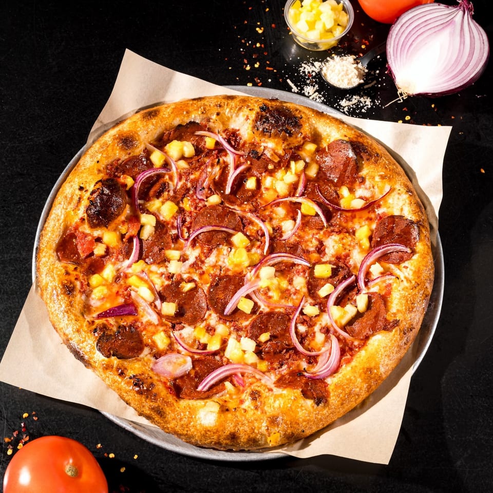 Vegan pizza with pepperoni, pineapple, and red onion on a dark surface with ingredients.