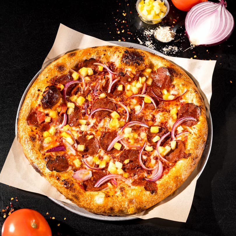 Vegan pizza with pepperoni, pineapple, and red onion on a dark surface with ingredients.
